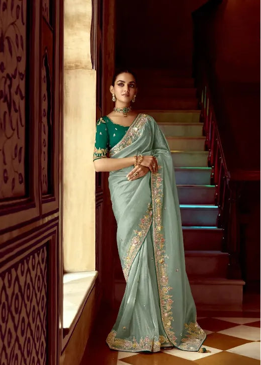 Woman in a light blue saree with green blouse standing in a decorated room.