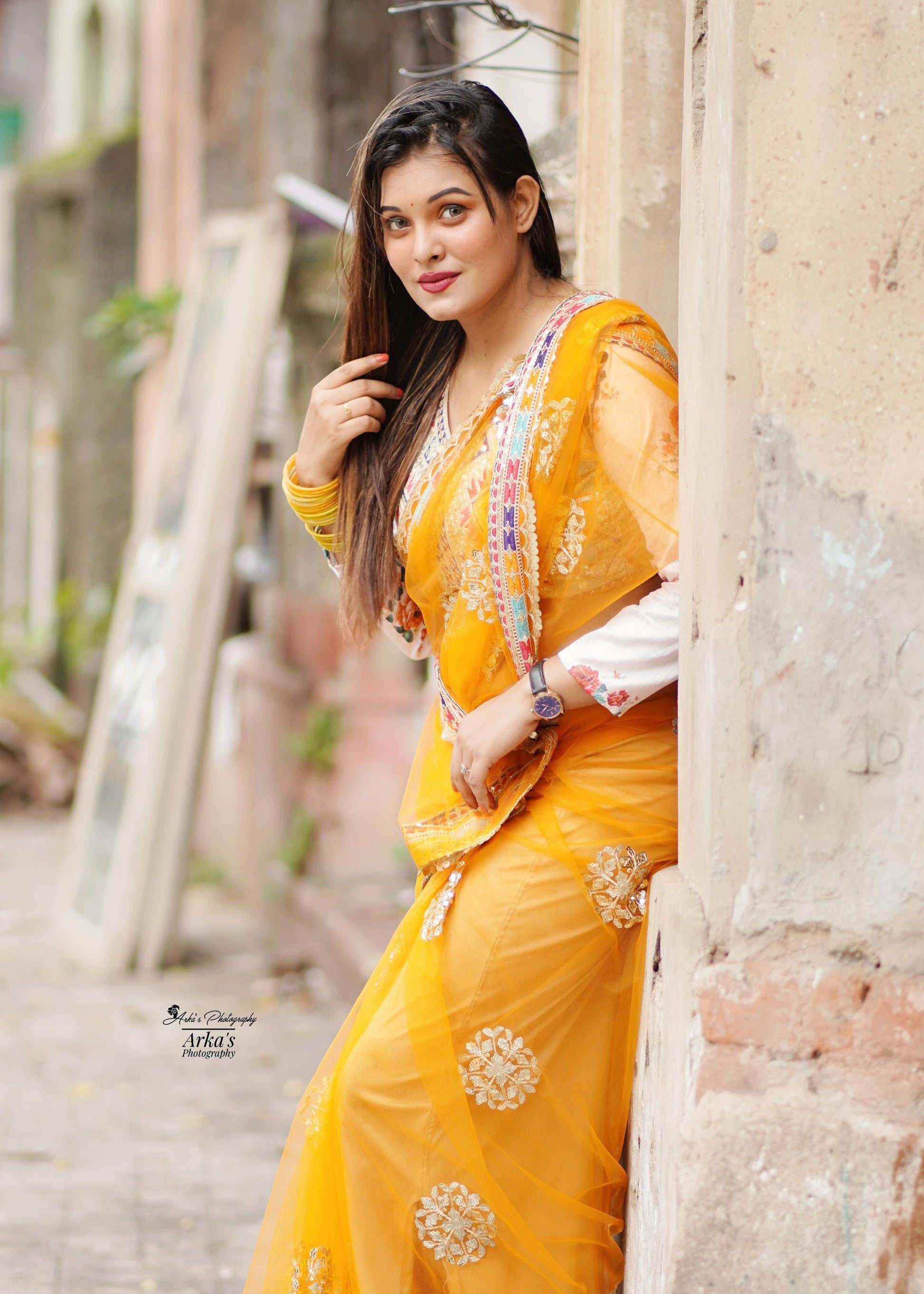 Woman in a yellow saree leaning against a wall in an urban setting