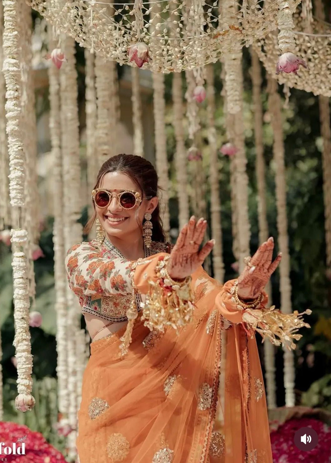 Woman in traditional outfit with floral decorations and sunglasses, surrounded by hanging floral arrangements.