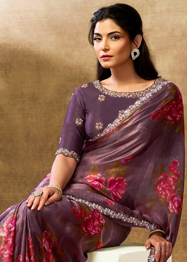Woman in a purple saree with floral patterns sitting against a beige wall.