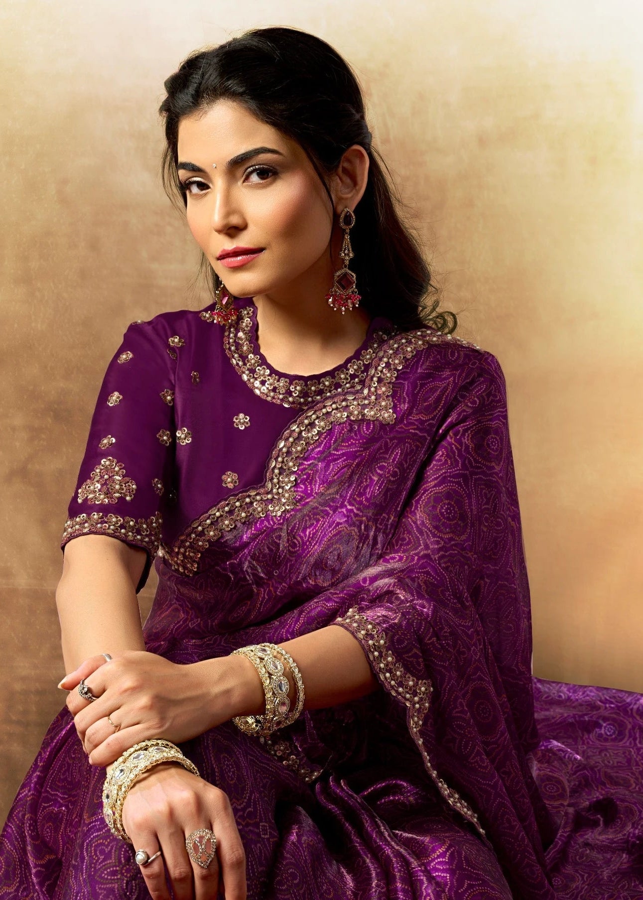 Woman in a purple traditional outfit with intricate designs, sitting against a beige wall.