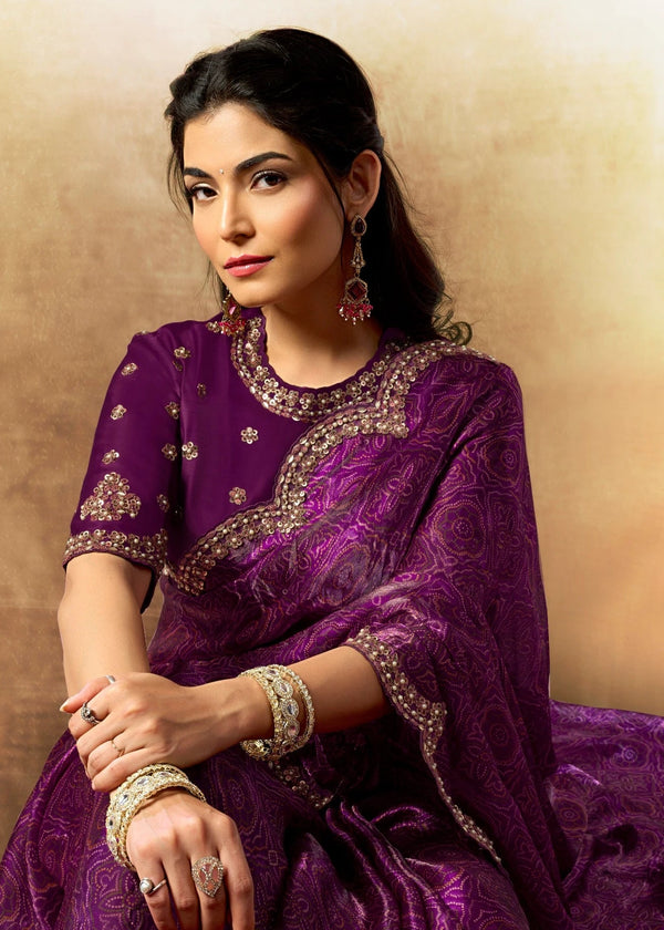 Woman in a purple traditional outfit with intricate designs, sitting against a beige wall.