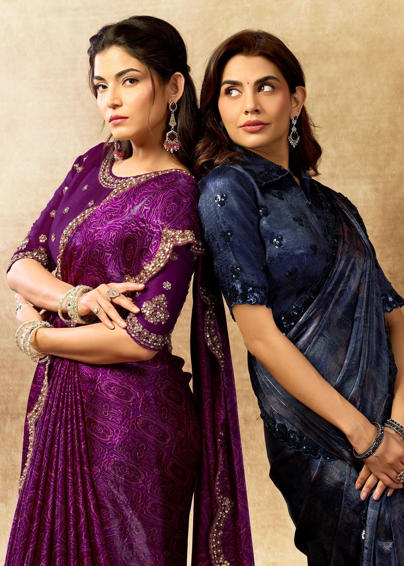 Two women in traditional attire standing next to decorative vases against a beige wall.