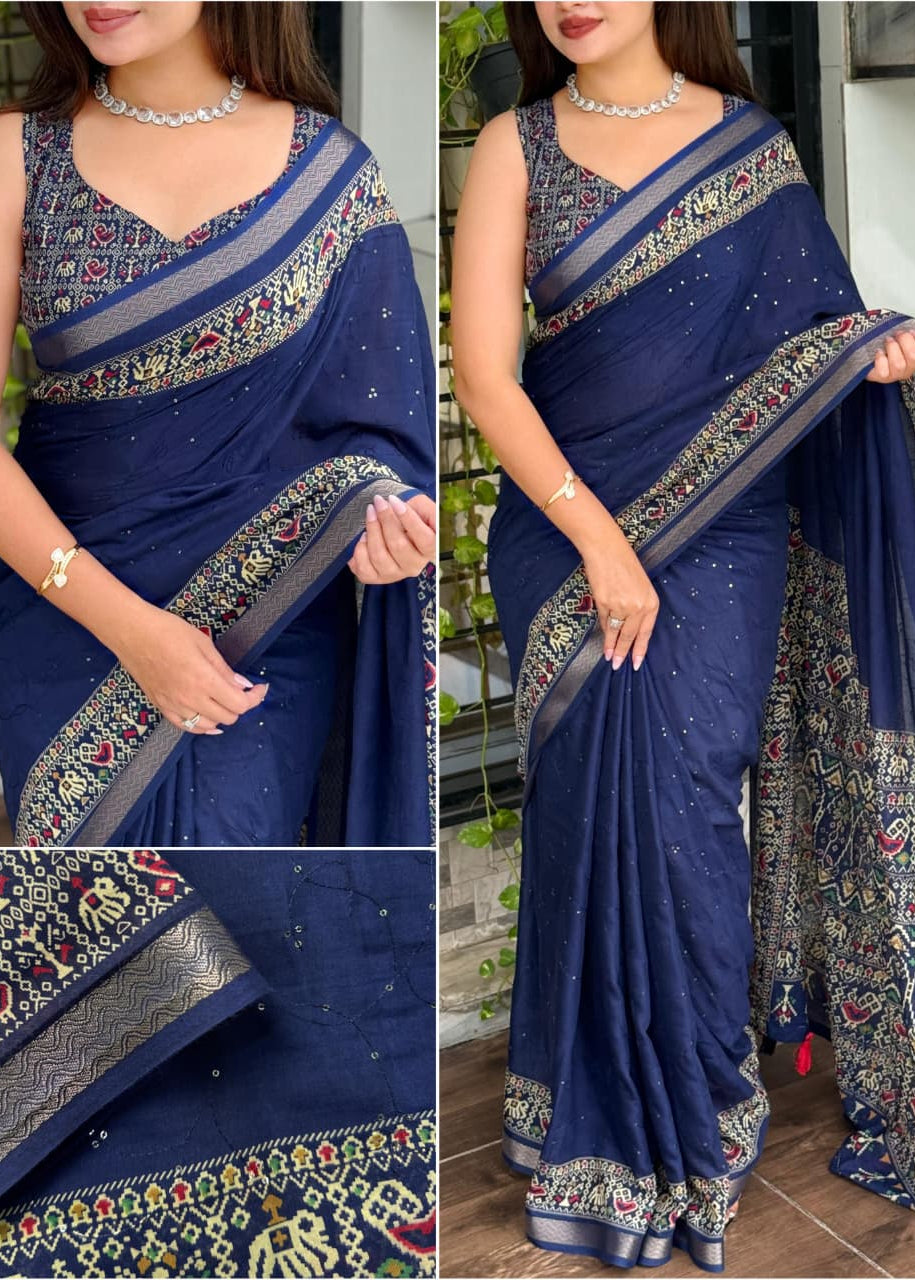 Woman wearing a blue saree with intricate patterns, standing outdoors.
