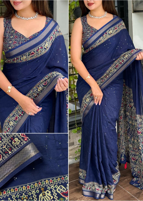 Woman wearing a blue saree with intricate patterns, standing outdoors.