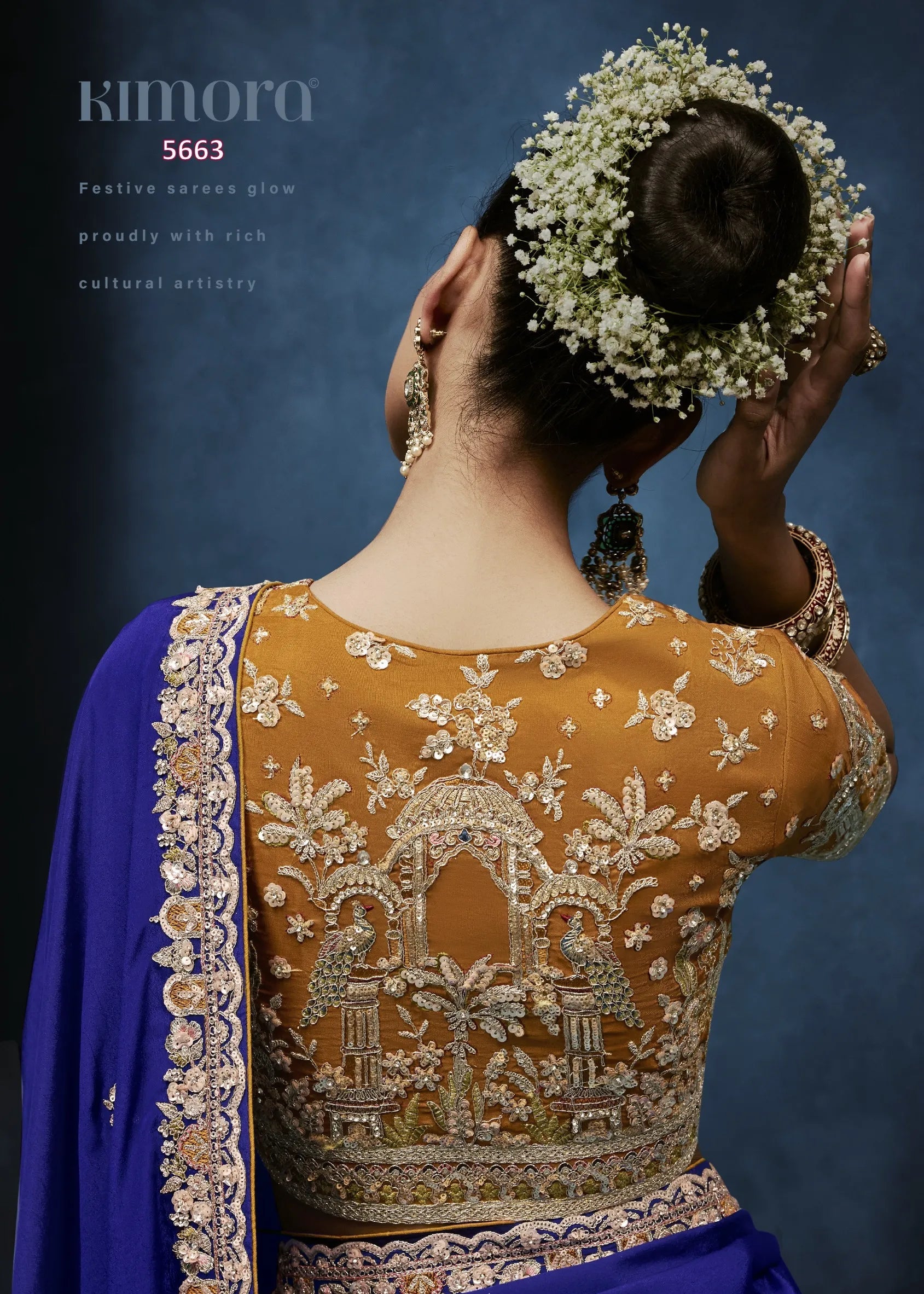 Woman wearing a traditional embroidered blouse with a blue saree, holding flowers in her hair against a dark background.