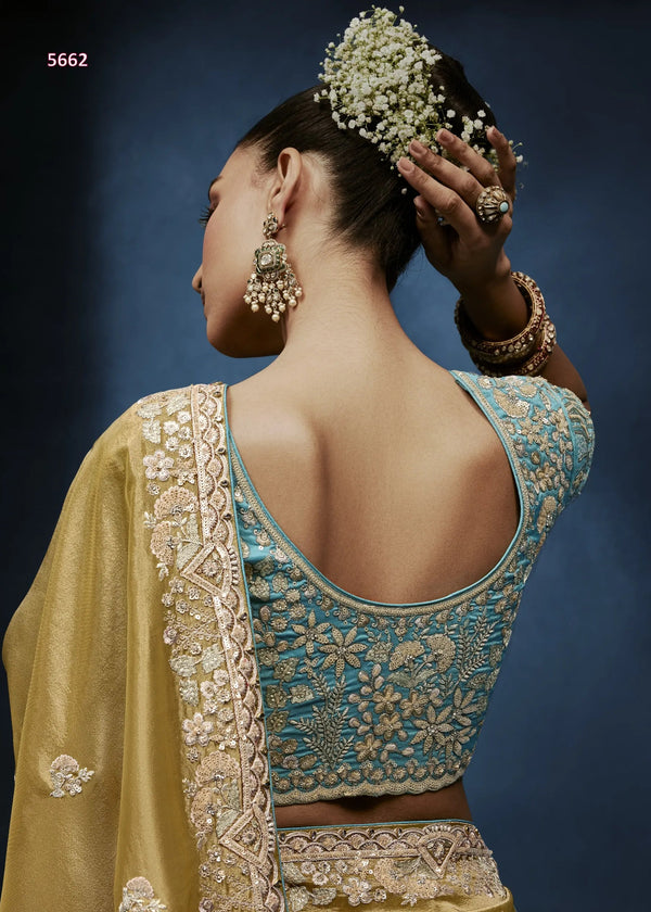 Woman wearing a traditional embroidered outfit with a blue blouse and yellow saree against a dark background.