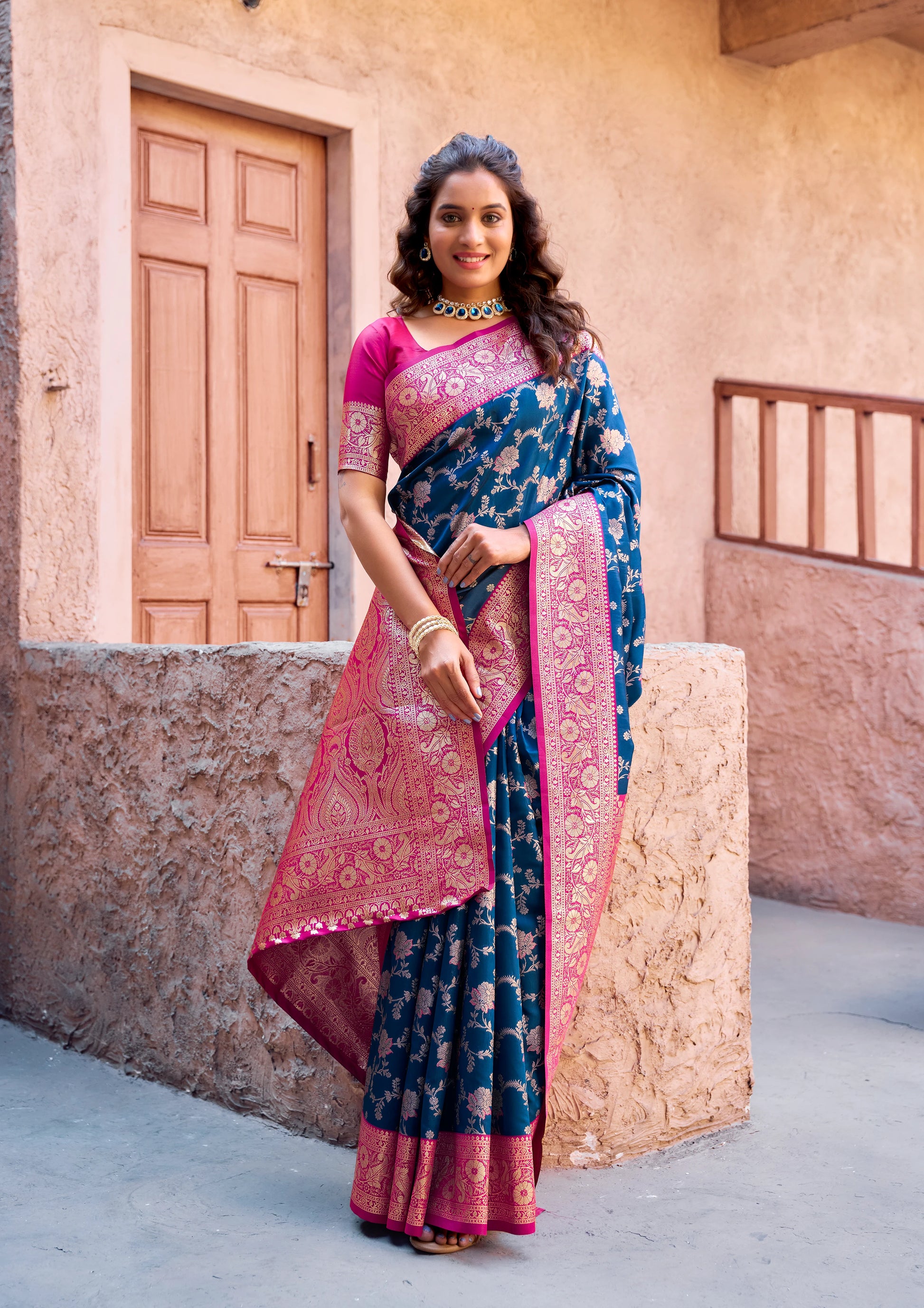 Woman in a traditional saree standing in front of a rustic building.