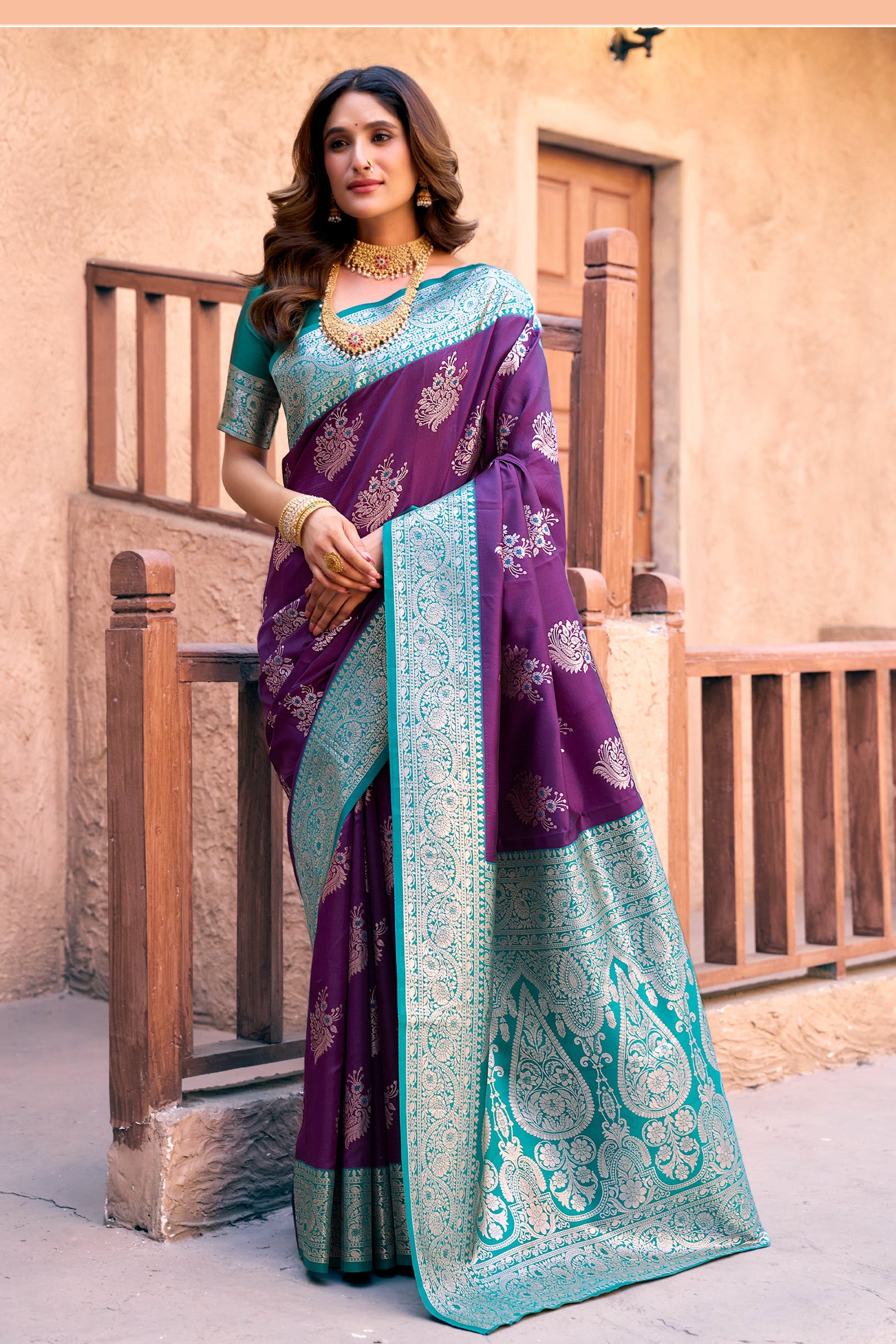 Woman wearing a purple and teal saree with a decorative pattern, standing in front of a beige wall.