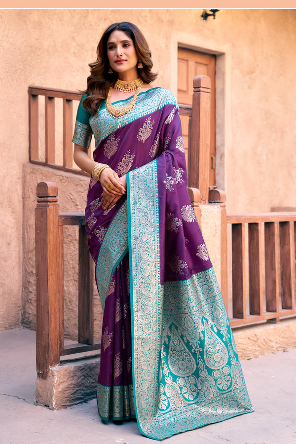 Woman wearing a purple and teal saree with a decorative pattern, standing in front of a beige wall.