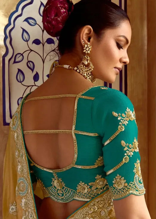 Woman in a teal blouse with gold embroidery and yellow saree, wearing jewelry.