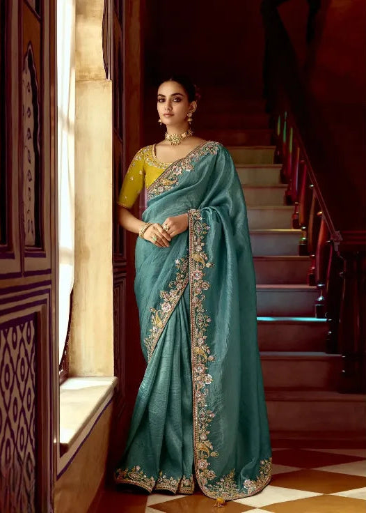 Woman in a teal saree with gold embroidery standing in an elegant interior setting.
