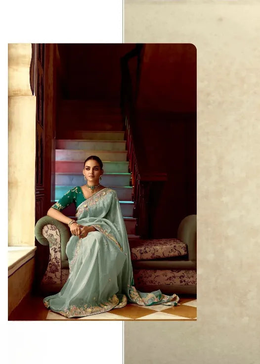 Woman in a green saree with intricate designs, sitting in a room.