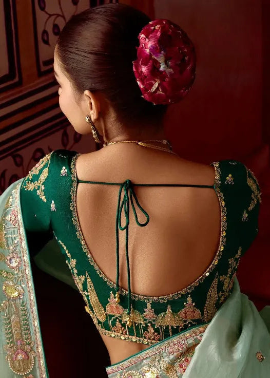 Woman in a green saree with intricate designs, sitting in a room.