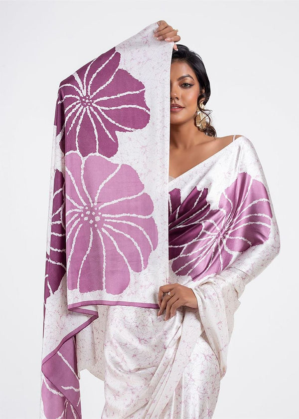 Woman wearing a white saree with purple floral patterns on a plain background