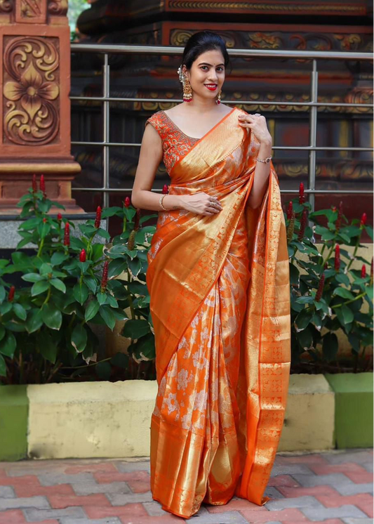 Woman in an orange saree standing in front of a decorative wall with plants.